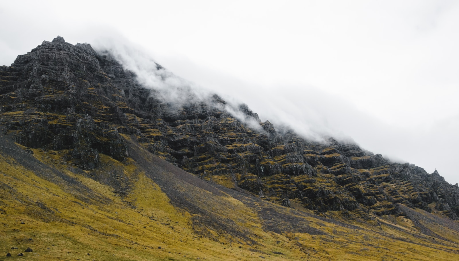 A fog line hugging a dark Icelandic mountain above a grassy slope.