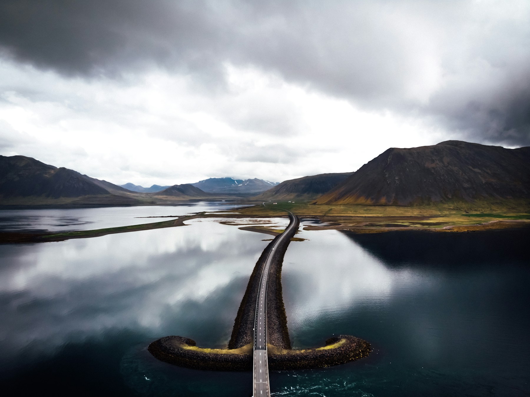 A road slicing through reflective water beneath dark Icelandic mountains.